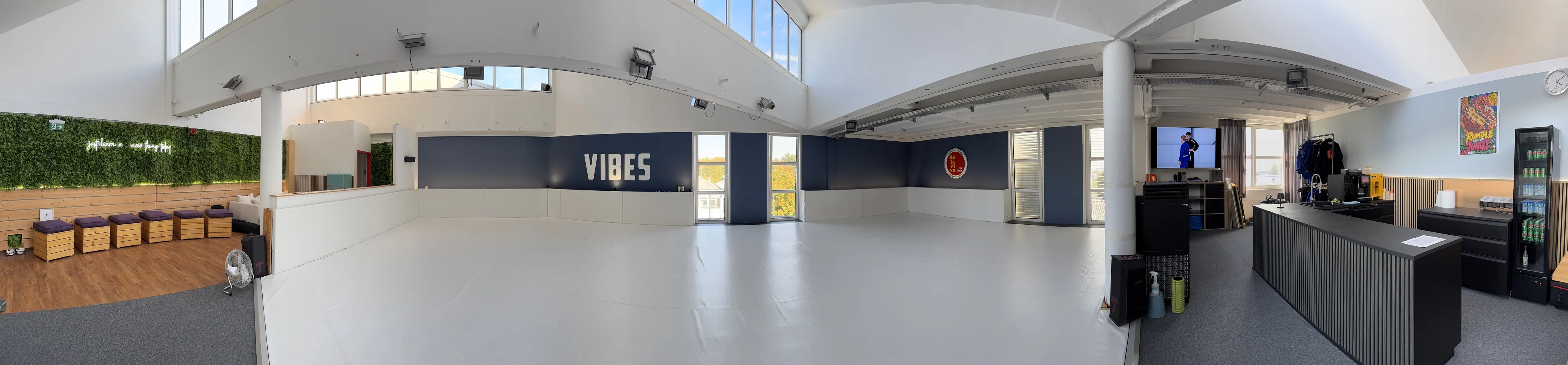 Panorama des VIBES-Studios: links der Sitzbereich mit grüner Wand, in der Mitte die helle Mattenfläche mit VIBES-Schriftzug, rechts der Empfang.